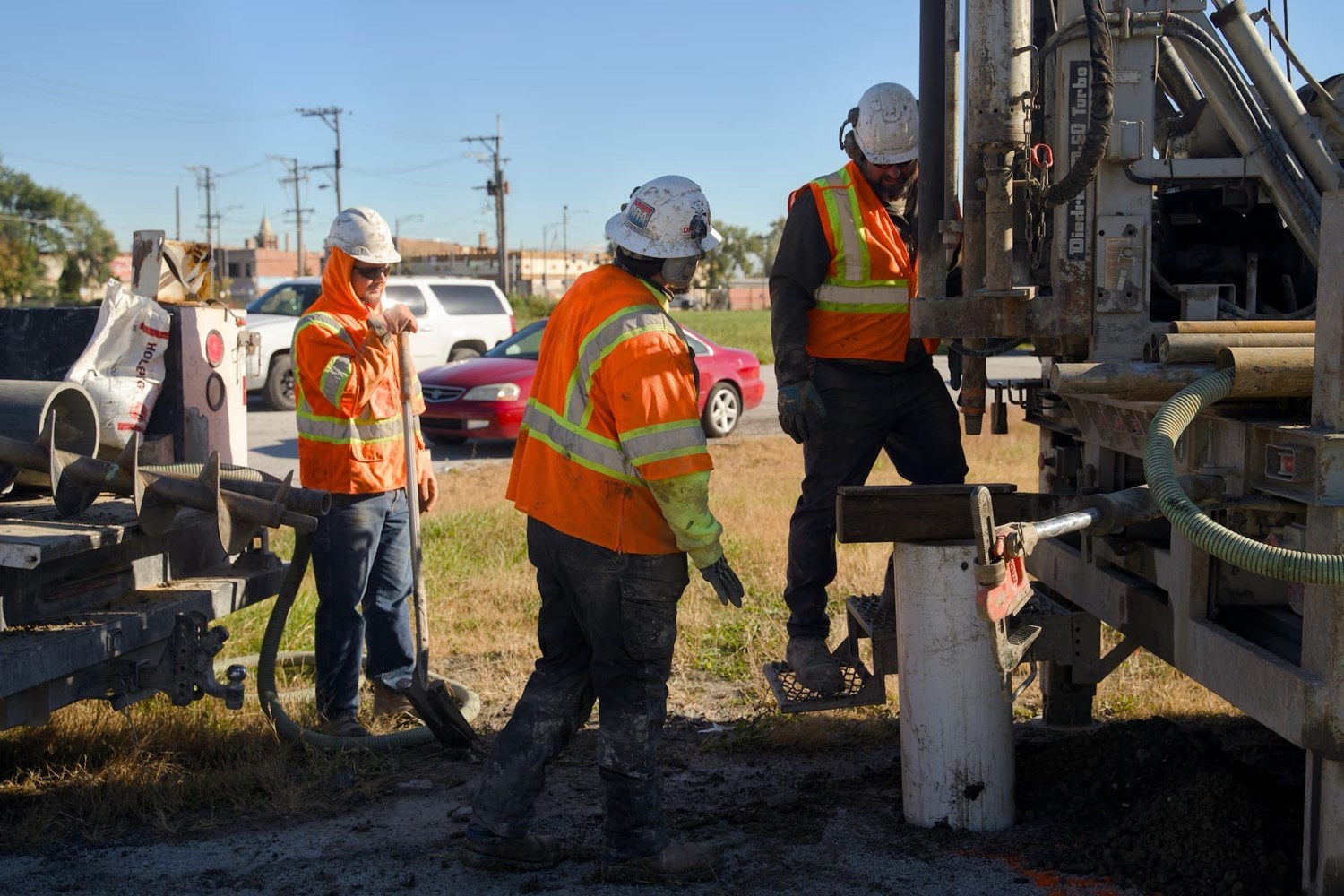 Three people in high visibility orange construction vests use a large piece of equipment for soil boring.