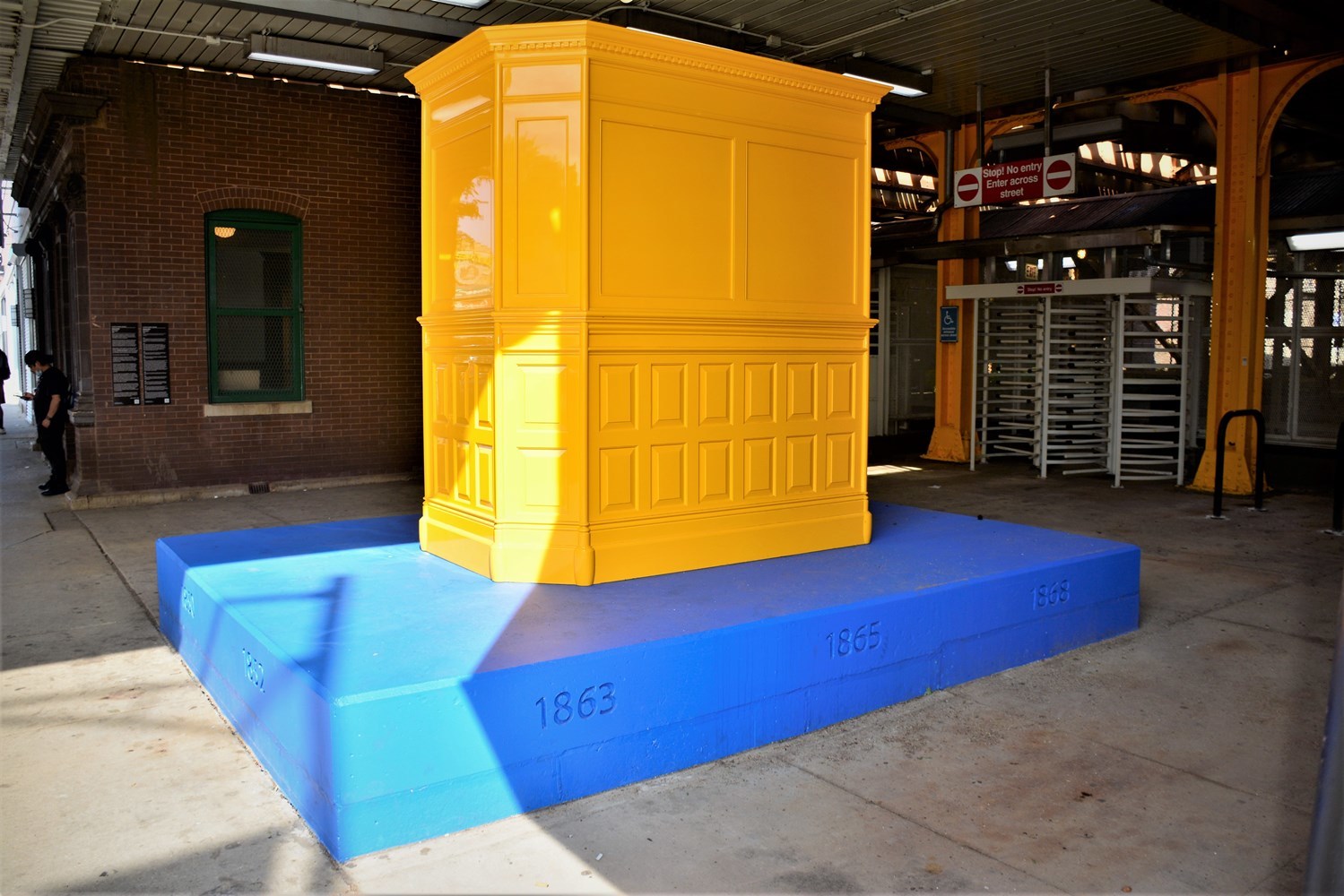A solid yellow replica of historic ticket agenct booth featured at Diversey sits in the plaza of the station and is just one four elements to the new art installation known as "Ordinary Relic."