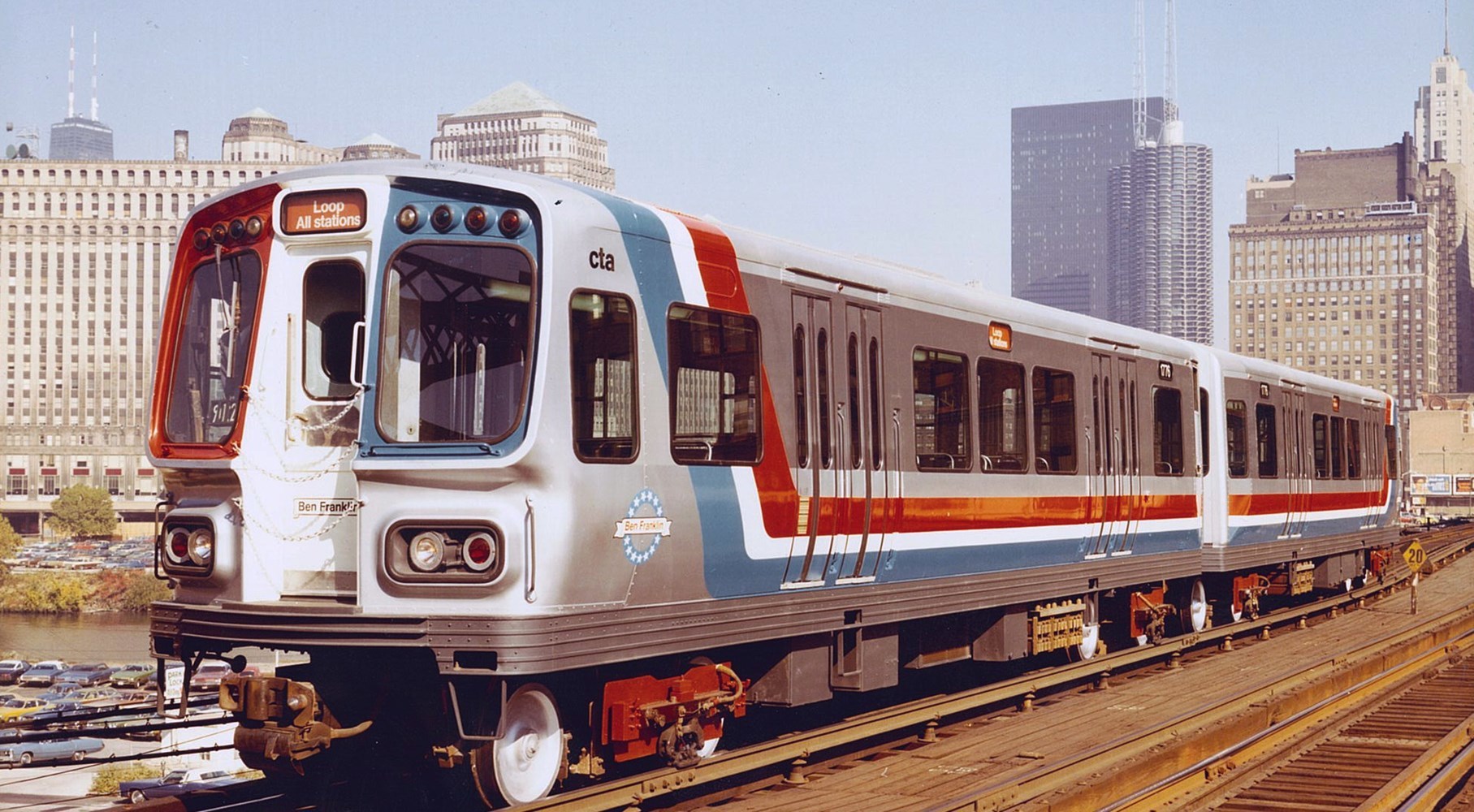2000-series cars are seen on the Lake Street Bridge in front of the Merchandise Mart, Hancock tower and IBM building in bicentennial livery honoring Ben Franklin