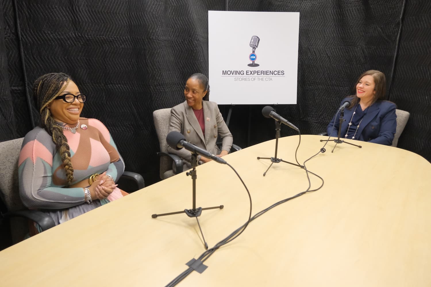 Three people recording at podcast. They are sitting a table with microphones. The walls behind them are covered in black curtains to dampen sound.